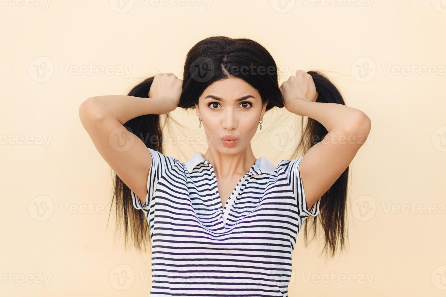 Portrait of pretty brunette female model makes two pony tails from her luxurious hair, has dark eyes, keeps lips round, wears casual t shirt, isolated over light studio background. People and fashion photo