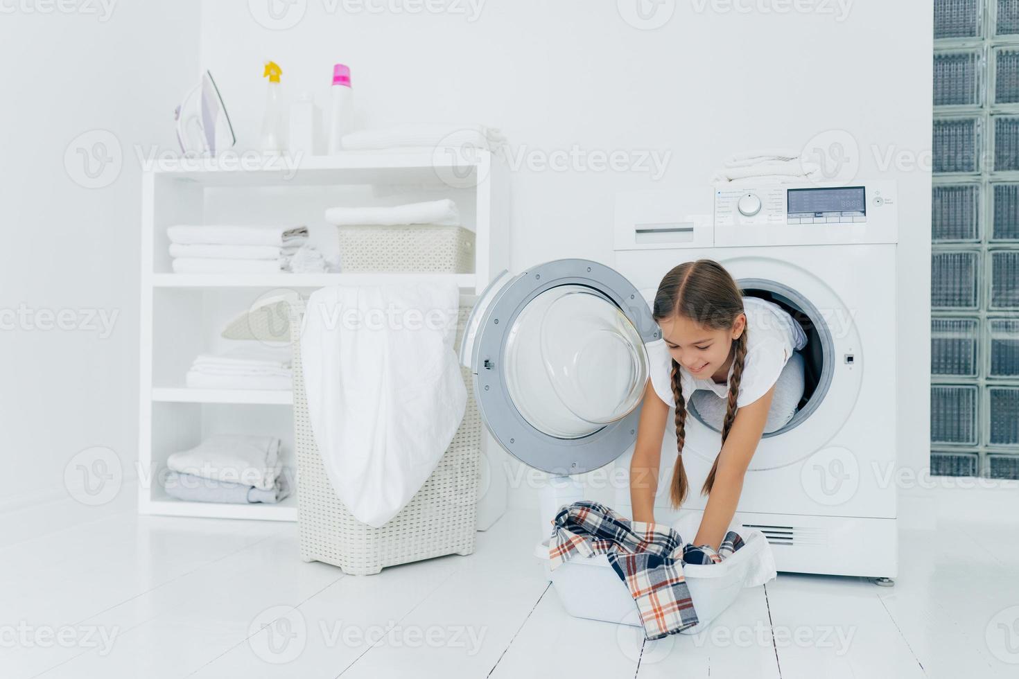 Pretty little girl householder poses inside of washing machine, takes checkered shirt from basin, engaged in laundry, has glad expression, two combed plaits. Childhood and washing day concept photo
