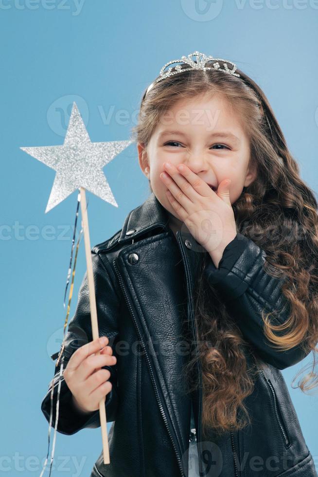 Overemotive positive small child covers mouth with one palm, wears fashionable black leather jacket and crown, holds magic wand in hand, isolated over blue background. Female kid poses indoor photo