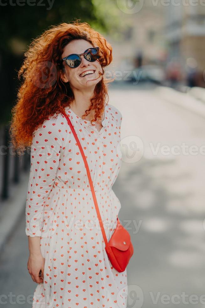 Optimistic foxy woman with glad expression, looks into distance, wears shades, long dress, strolls outdoor on street, enjoys nice summer day, has broad smile. People, lifestyle, rest and lifestyle photo