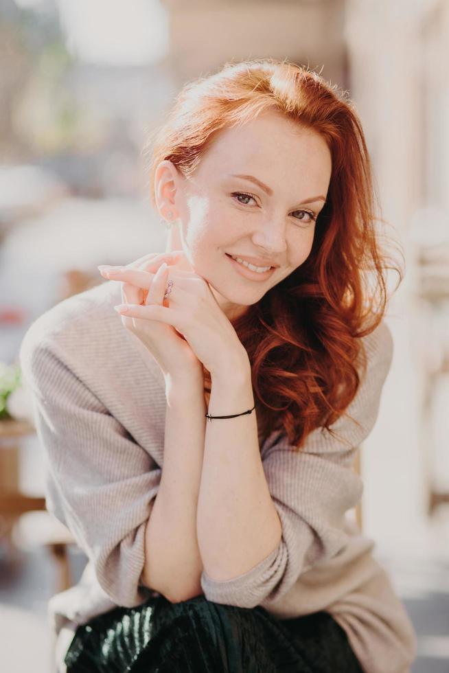 Image of lovely redhead female model smiles happily, has healthy skin, make up, keeps hands together, dressed in oversized jumper, poses outside, smiles gladfully. Having nice day or weekend photo