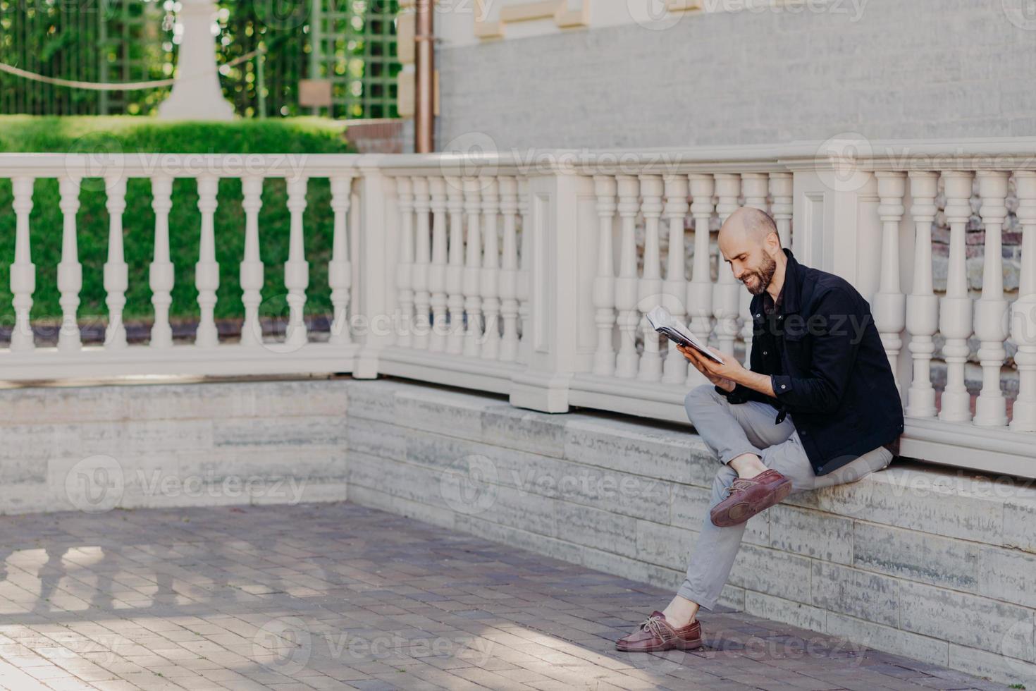 People, positive emotions and hobby concept. Unshaven bald middle aged man with happy expression, focused into book, reads something interesting, sits crossed legs, outdoor, has spare time. photo