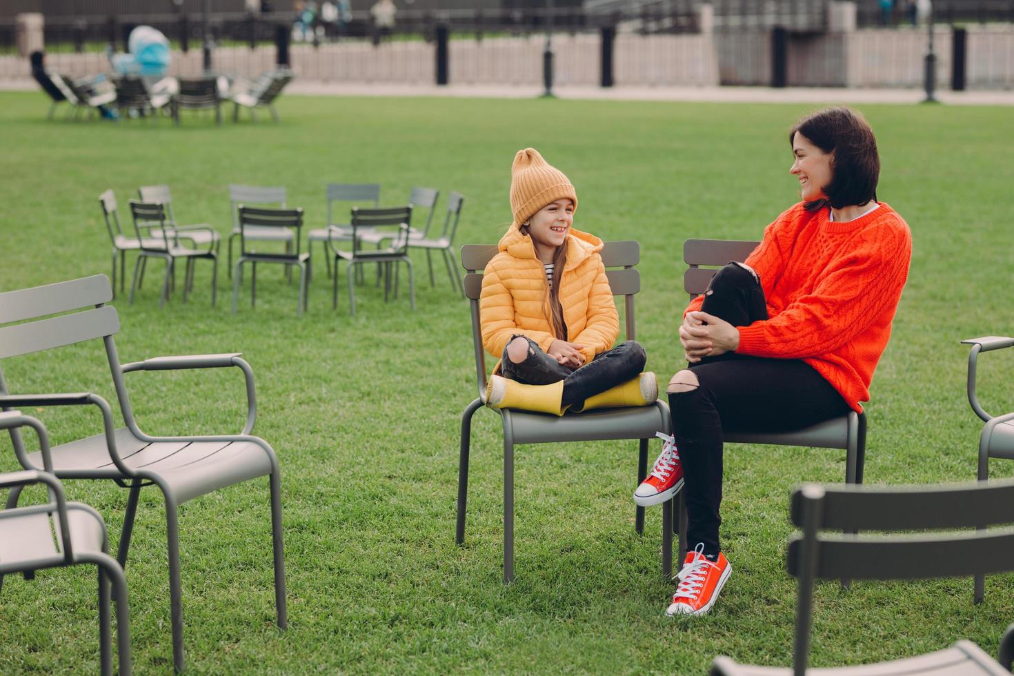 foto de una joven madre e hija positivas y amigables posan afuera en la zona de recreación con césped verde en sillas, vestidas con ropa de moda, se ríen de algo divertido. familia y comunicacion