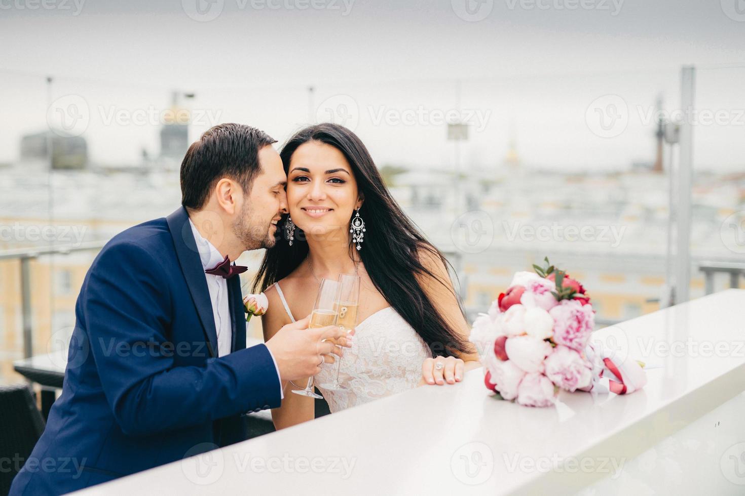 los recién casados románticos beben champán, tintinean copas, están muy felices de celebrar su boda, sienten pasión y gran amor entre ellos. bella novia recibe un beso del novio foto