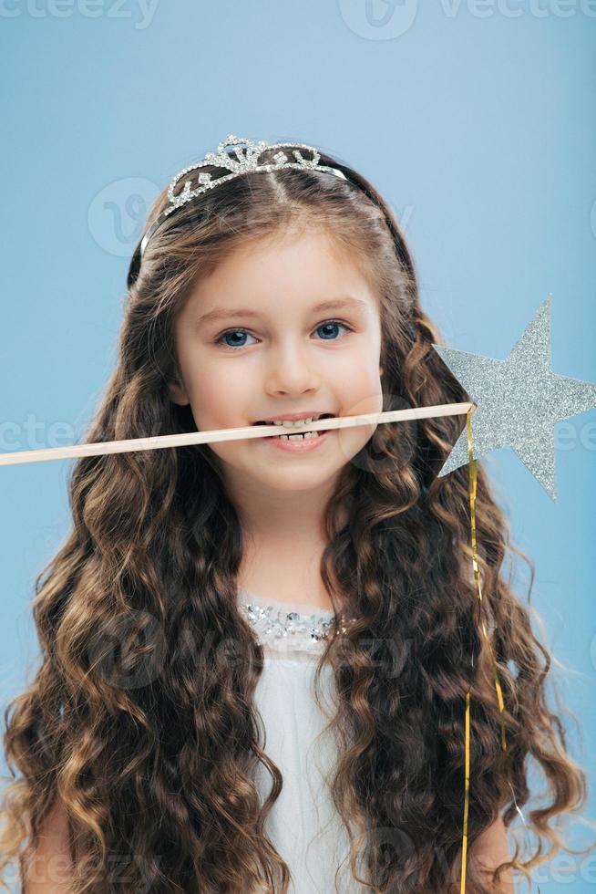 Photo of attractive woman kid with pleasant appearance, has long dark hair, holds magic wand in mouth, poses over blue background, dressed in princess dress. Vertical image. Childhood concept