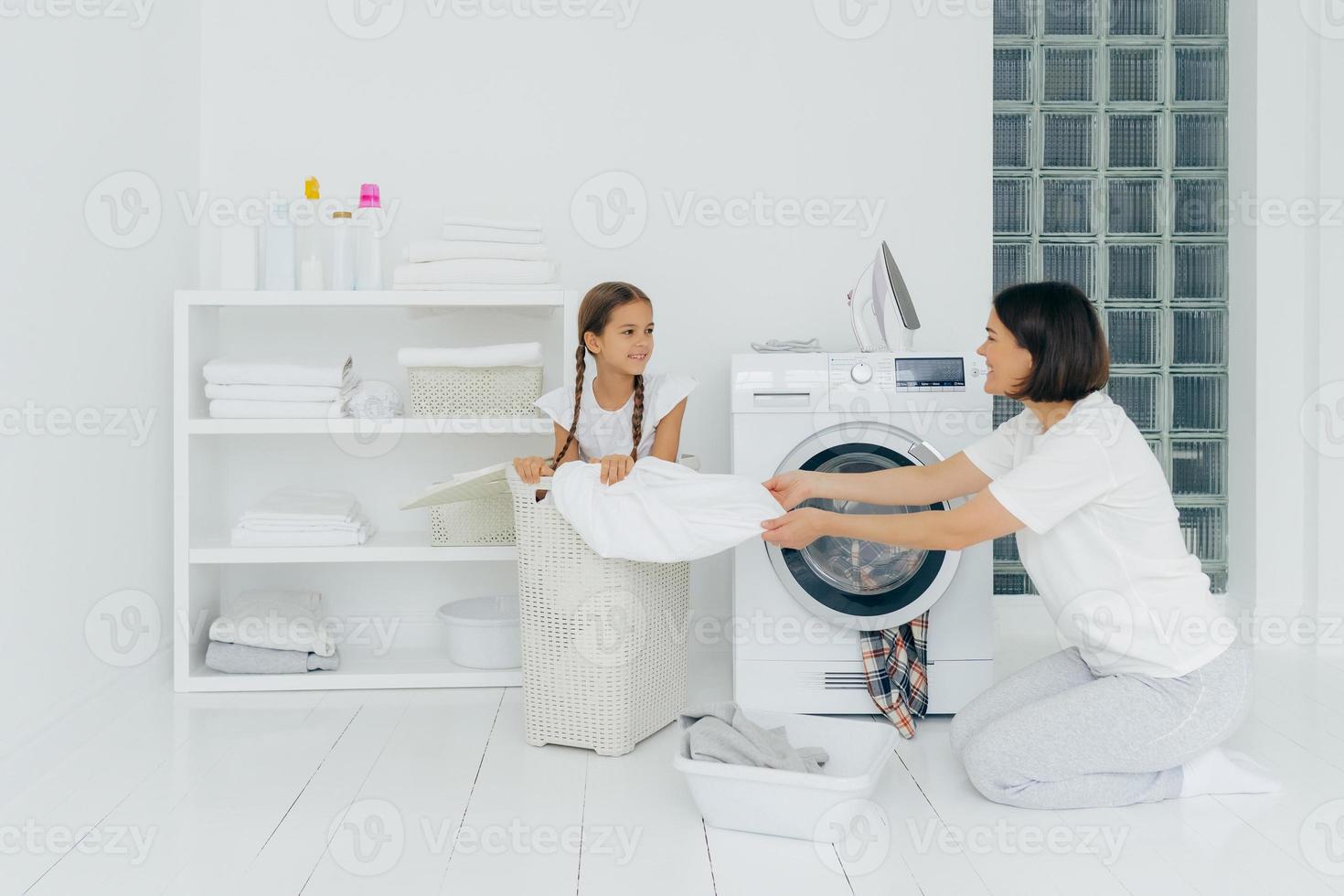 el niño pequeño ayuda a mamá a lavarse, se sienta en una cesta con ropa sucia. el ama de casa ocupada hace las tareas del hogar, lava la ropa en la lavadora, carga la lavadora, posa en un espacioso baño blanco. concepto de hogar foto