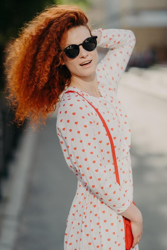 Vertical shot of lovely woman in trendy sunglasses, has curly bushy red hair, carries small bag, poses outdoor over blurred background, has stroll in city, enjoys summer holidays, explores sights photo