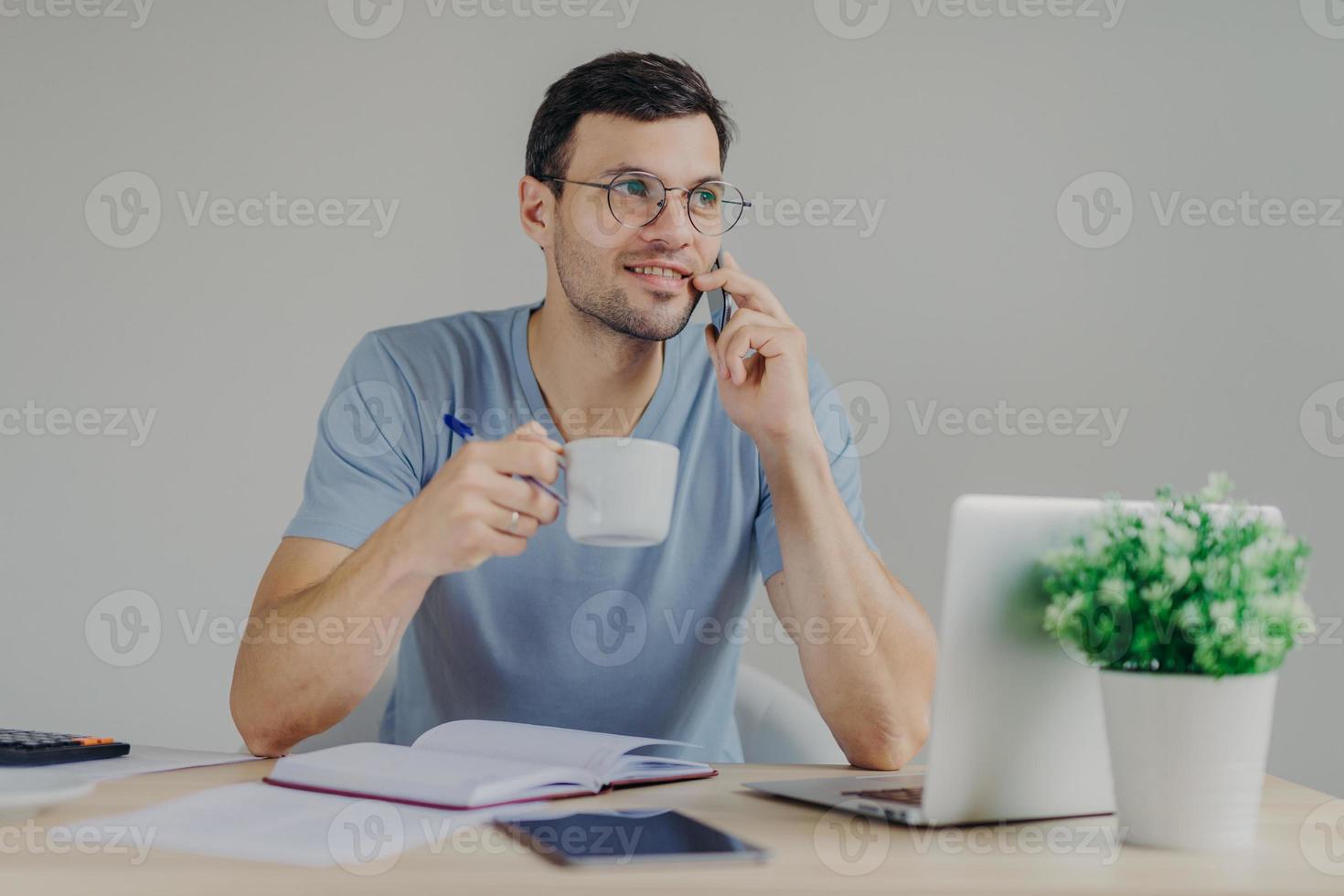 un contador de aspecto agradable con gafas redondas toma una bebida caliente, tiene una conversación móvil, administra las finanzas, trabaja con una computadora portátil, escribe notas en el diario. el hombre responde a la llamada del banco. foto