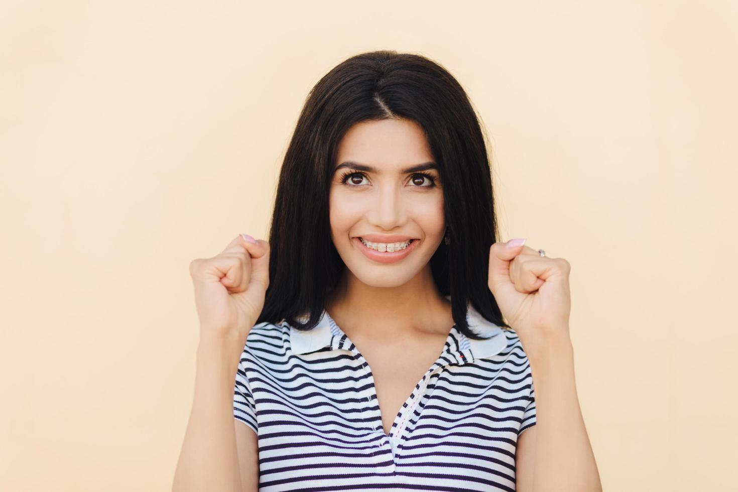 Headshot of happy brunette female model clenches fists, hopes for something good, has broad smile with white perfect teeth, celebrates success, isolated over beige studio wall. I will do it photo