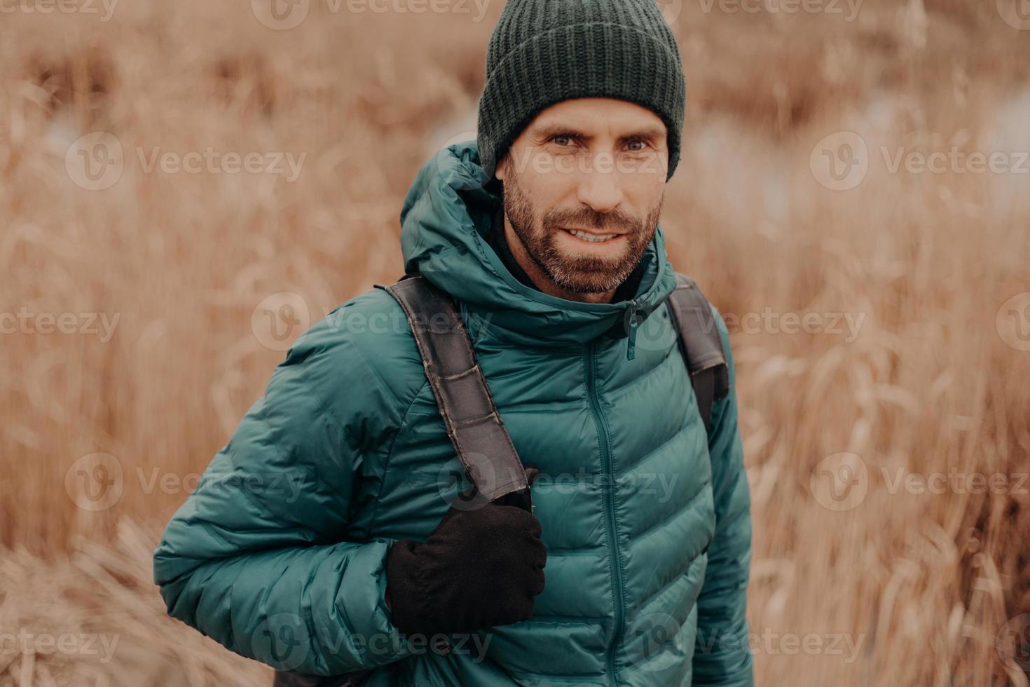 Headshot of atrractive blue eyed man has dark stubble, wears hat, jacket, gloves, carries rucksack, has outdoor stroll, models against blurred background. People, leisure, traveling and rest concept photo
