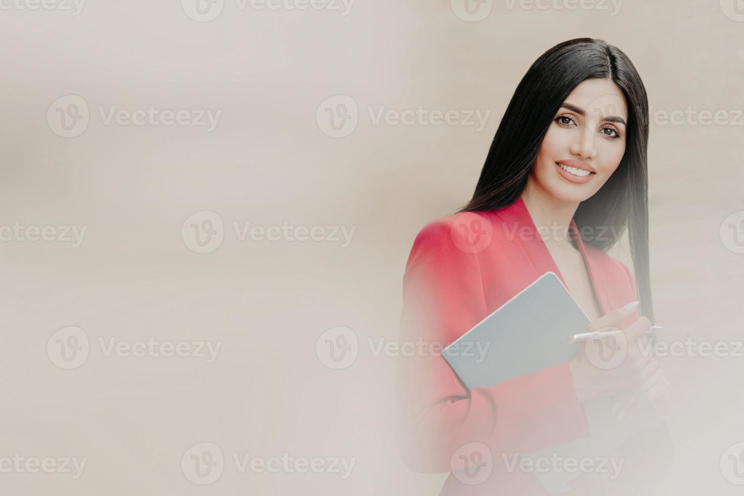 Horizontal shot of lovely brunette woman with happy facial expression, dressed in formal clothes, holds notebook and pen, has black straight hair, stands over white blurred background with free space photo