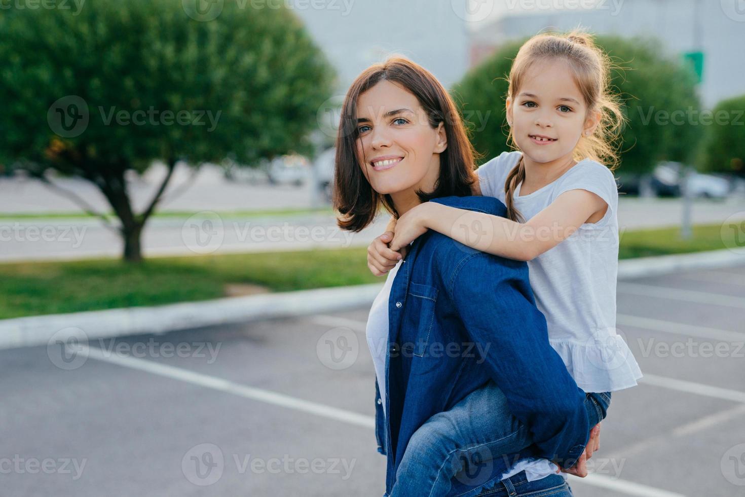 Outdoor shot of smiling woman gives piggyback to her daughter, have walk in open air, smile positively, play and have fun together, pose against asphalt and green trees background. Family concept photo