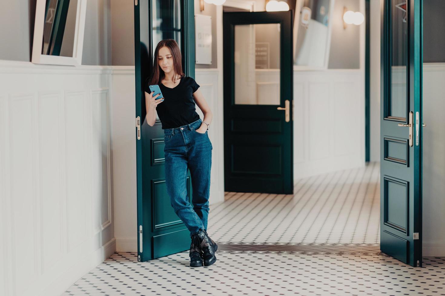 Full length shot of serious fashionable woman concentrated into cell phone, wears black t shirt, jeans and boots, reads information in internet, stands indoor near doors connected to wireless internet photo