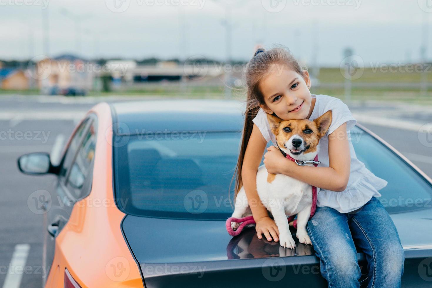 Small attractive female child embraces her favourite dog, sit together at trunk of car, have rest after stroll, enjoy summer day, have friendly relationship. Children, pets and lifestyle concept. photo