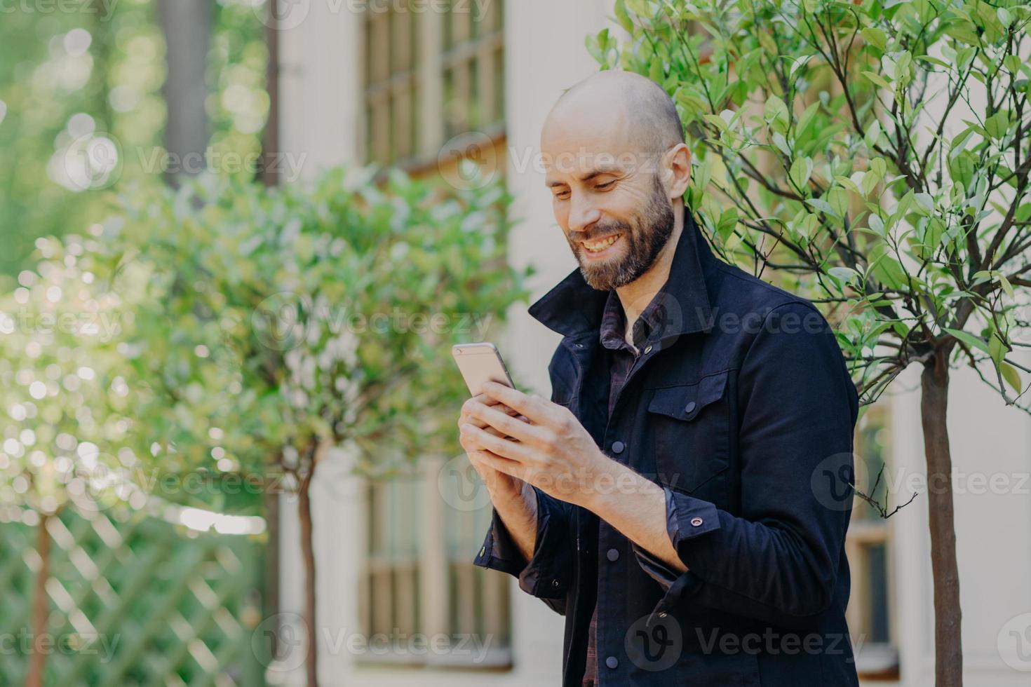 Positive bald young male with stubble, holds modern cell phone, enjoys online communication in social networks, has glad expression, dressed in black jacket, poses outdoor against green trees photo