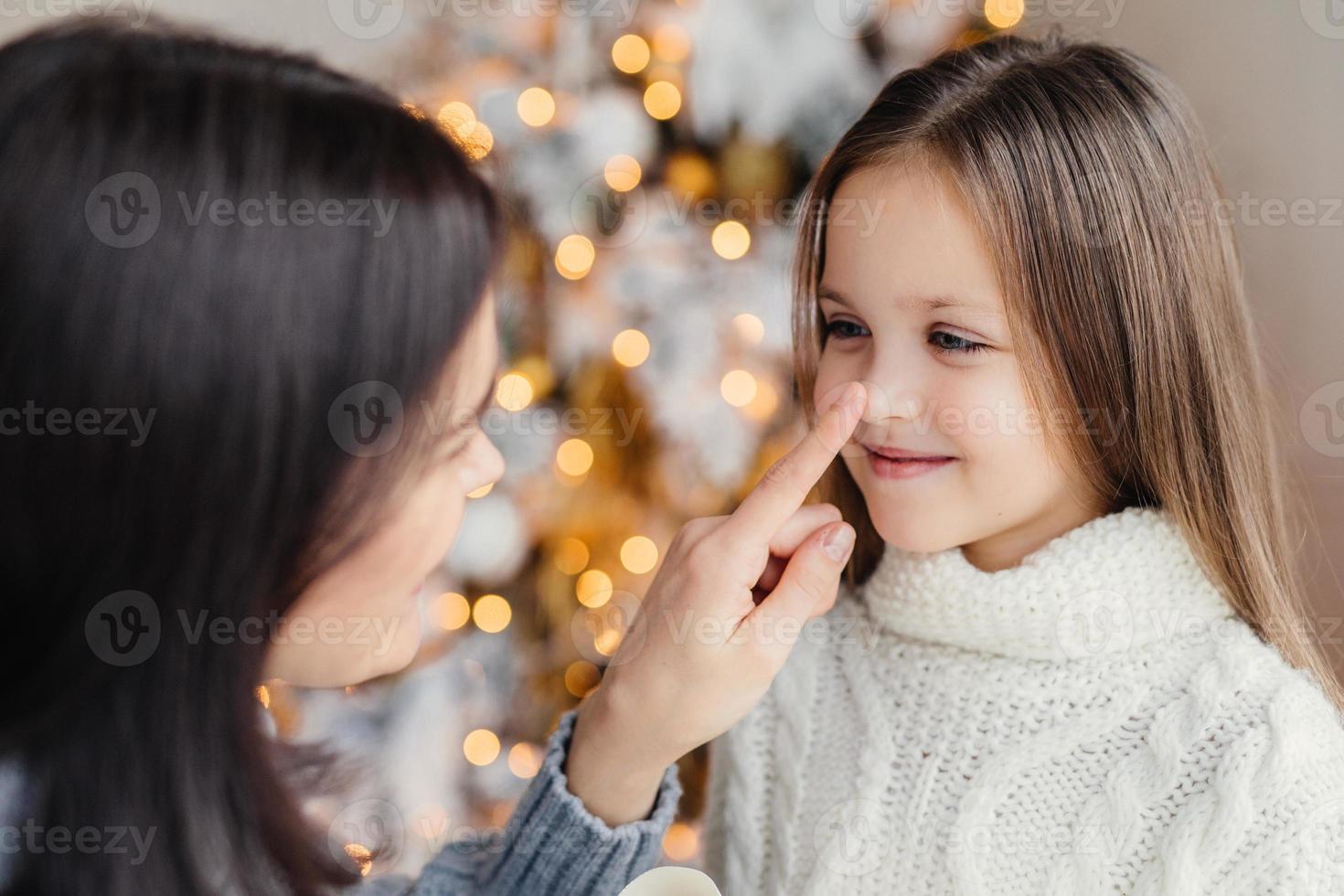 Close up portrait of beautiful little girl with long hair, has fun with her mother, looks in eyes, stand together against decorated fir tree with garlands and lights background. Celebration concept photo
