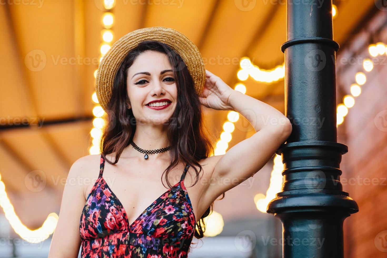 mujer elegante de moda con vestido con estampado de flores y sombrero de paja, posando en la cámara con una sonrisa, sintiéndose relajada, con buen humor. gente, belleza, estilo de vida, expresión facial, concepto de juventud foto