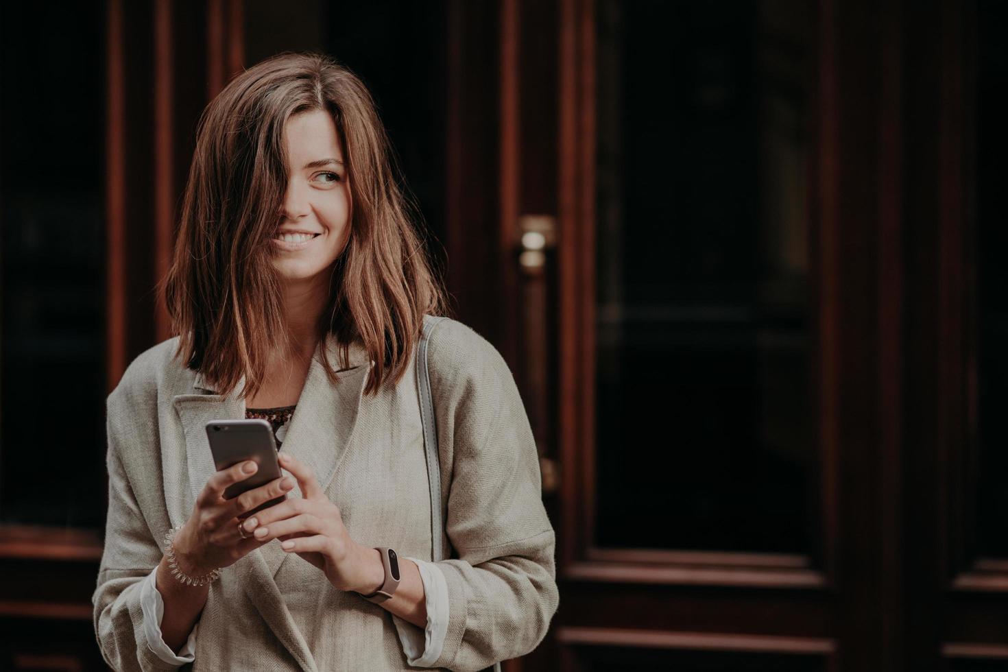 Emotional delighted happy brunette young woman with dark hair, uses mobile phone for texting messages, dressed in elegant raincoat, stands near door, cant imagine life without modern technoliges photo