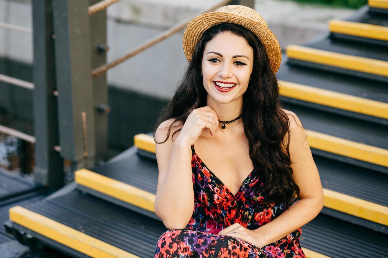 mujer sonriente con cabello oscuro y lujoso vestido y sombrero de paja sentada en las escaleras mirando con una mirada encantadora a la distancia. mujer joven tranquila con apariencia atractiva sentada al aire libre foto