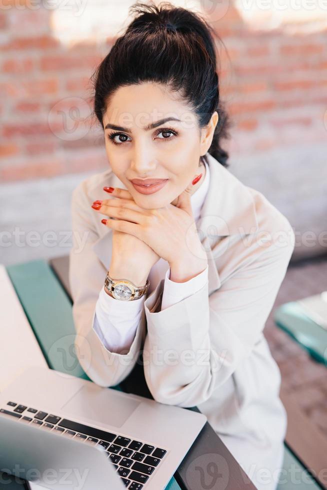 Young brunette businesswoman with charming eyes, gentle hands with red manicure wearing watch on hand and white coat holding hands under chin sitting near her laptop having rest after hard work photo