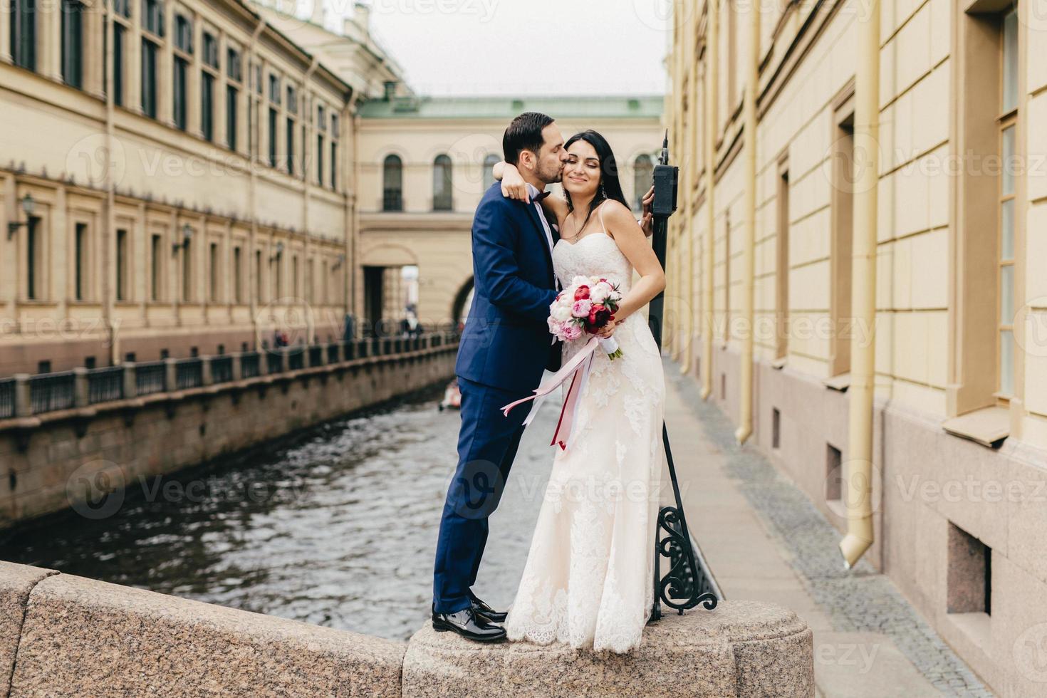 cariñosa pareja casada se besa apasionadamente mientras se para en el puente cerca del río en la ciudad antigua, celebra su boda, feliz de crear su familia amistosa. los recién casados románticos posan al aire libre foto