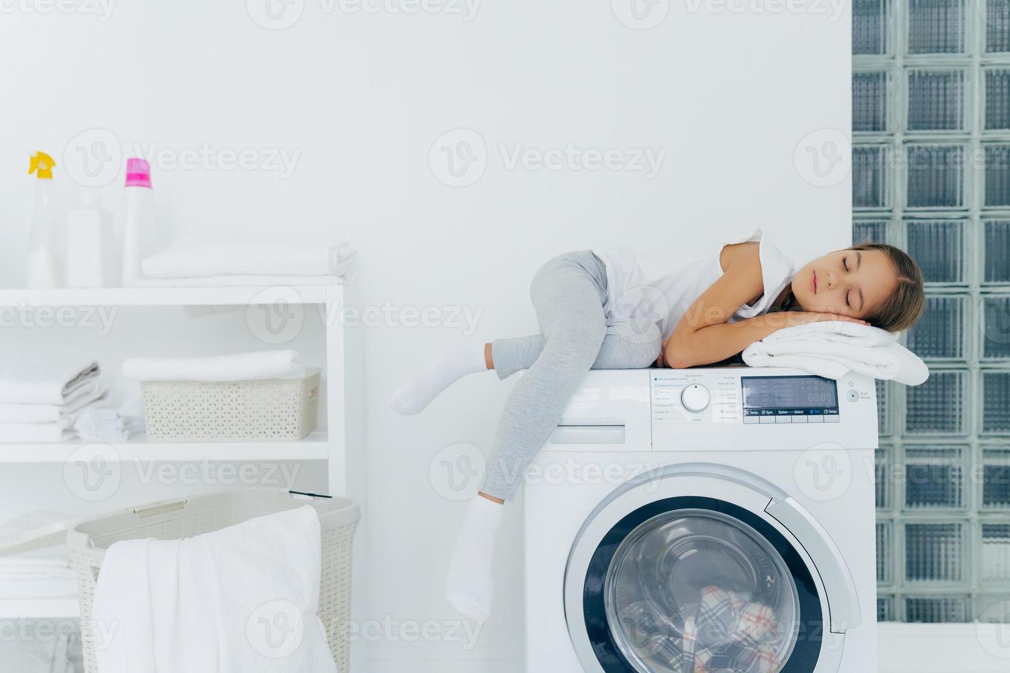 Horizontal shot of little girl has rest on washing machine, feels tired after helping mum about house, sleeps on washer poses in laundry room with console, detergents, linen around. Household routine photo