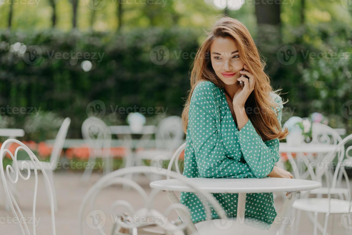 Outside shot of charming young woman with long hair, wears polka dot green shirt, sits at table in outdoor cafe, has pleasant talk via modern smartphone, has dreamy expreession. People and lifestyle photo