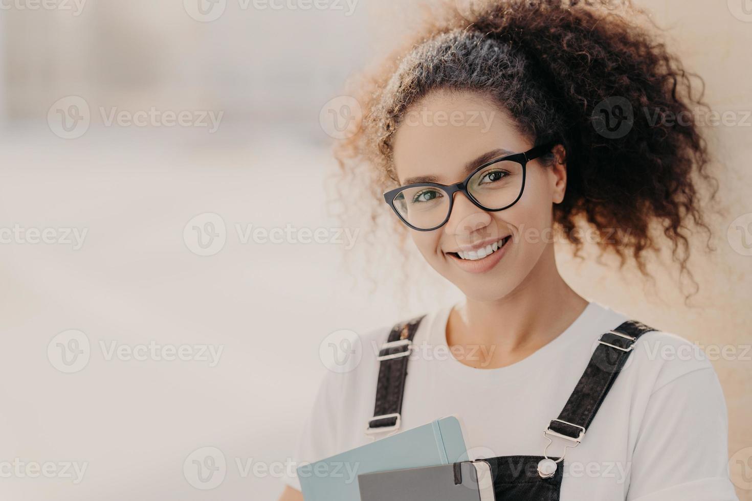 Horizontal shot of beautiful woman with crisp hair combed in pony tail, carries notepad, wears white t shirt and overalls, enjoys studying, poses against blurred background with empty space for text photo