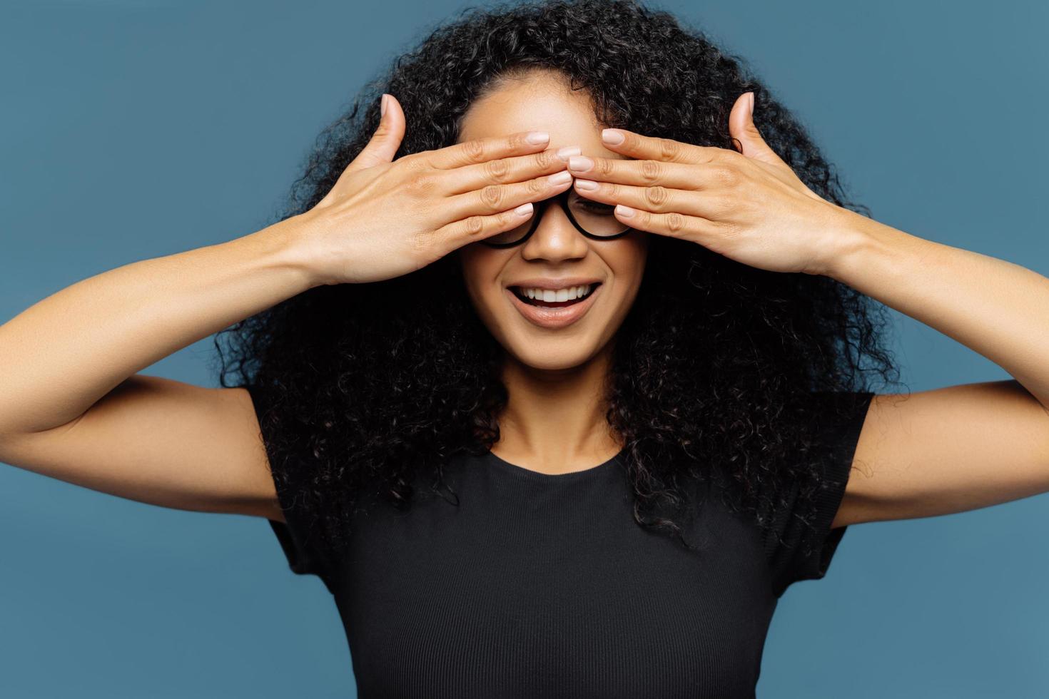 Secret woman with curly hair, covers eyes, wears spectacles, waits for surprise, dressed in black casual t shirt, poses against blue background, has manicure anticipates for miracle. Female hides face photo