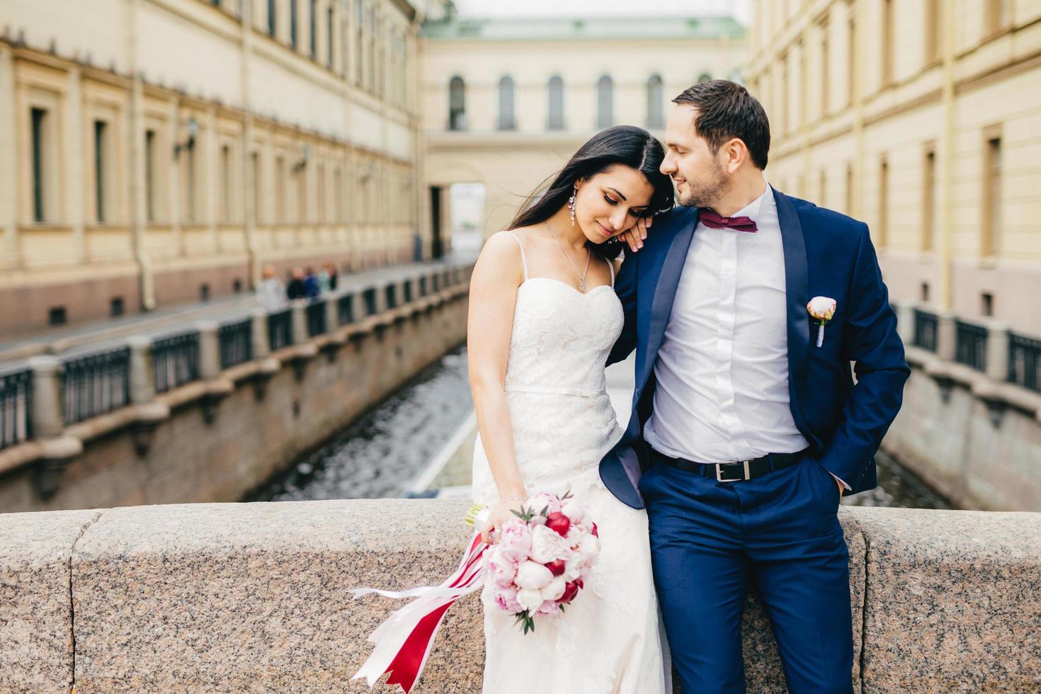 una mujer joven de buen aspecto con una apariencia atractiva viste un vestido de novia blanco, sostiene un bonito ramo, se apoya en el hombro de los amantes, disfruta de la unión y la atmósfera tranquila mientras se encuentra sola en el puente. foto