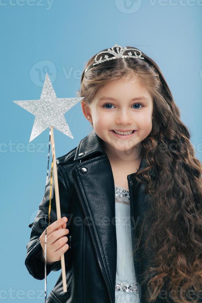 Satisfied small female child has long curly hair, dressed in black leather jacket, wears crown, holds magic wand, isolated over blue background. Dark haired positive kid anticipates for miracle photo