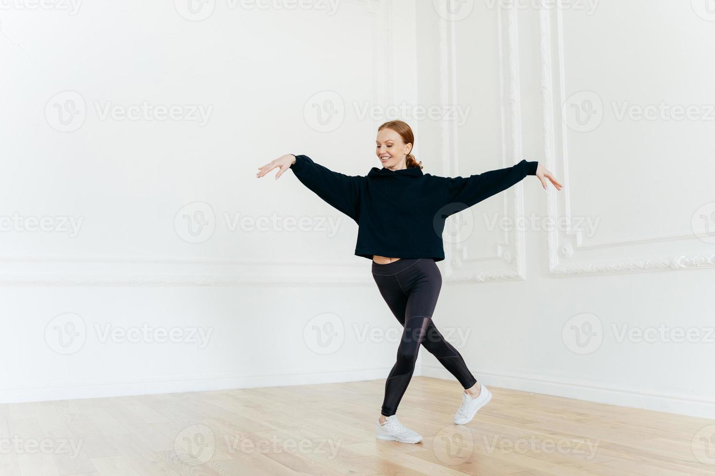 Professional female ballerina practices dance in hall, stretches hands, stands with crossed legs, has gentle smile on face, dressed in black sportswear, enjoys spare time for hobby. Healthy lifestyle photo