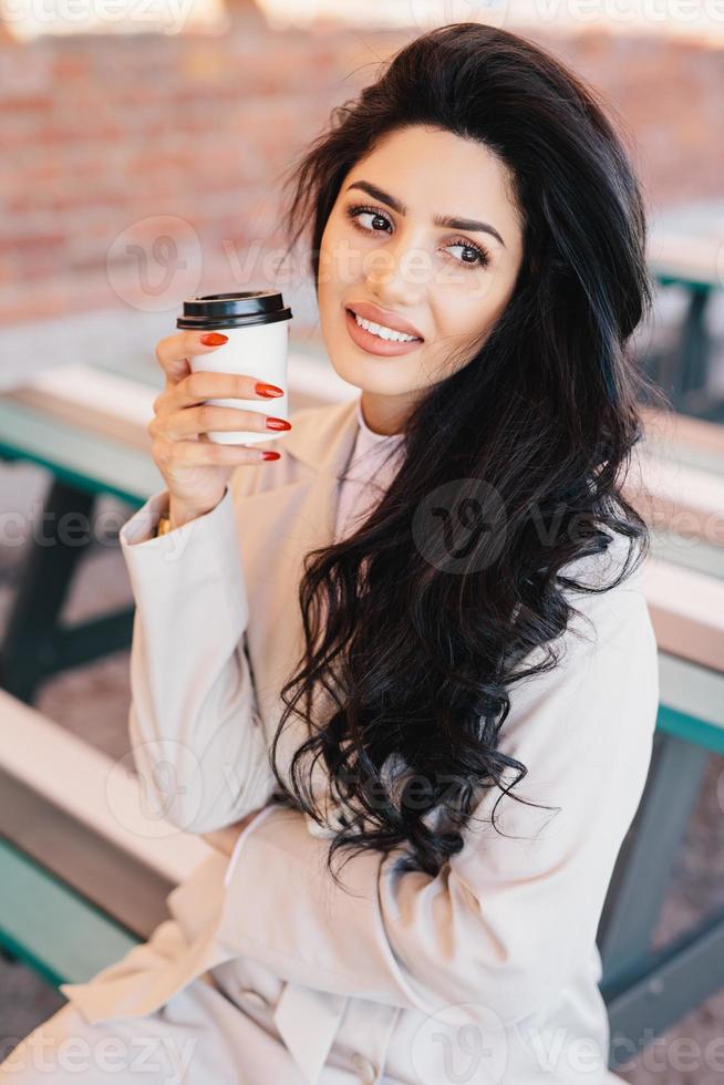 hermosa mujer con cabello oscuro, cejas y labios bien formados mirando a un lado y sonriendo bebiendo capuchino en una taza para llevar sentada en un banco al aire libre. hermosa mujer sonriente con buena relajación foto