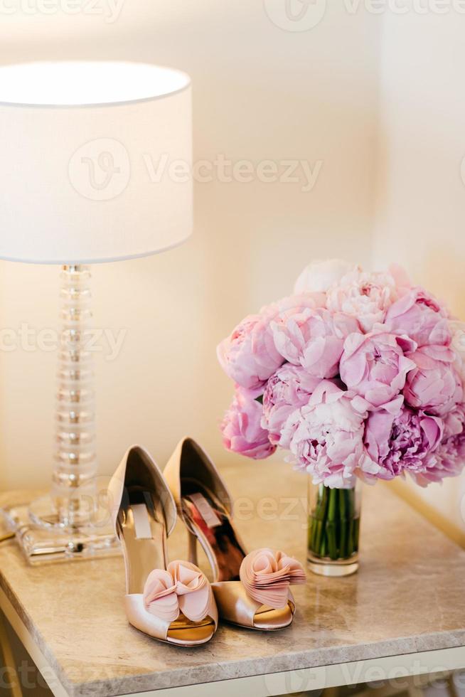 Beautiful shoes with high heels, lamp and bouquet with pink flowers standing on bedside-table. Bouquet and shoes of bride. Wedding shoes and flowers. Preparation for special occasion photo