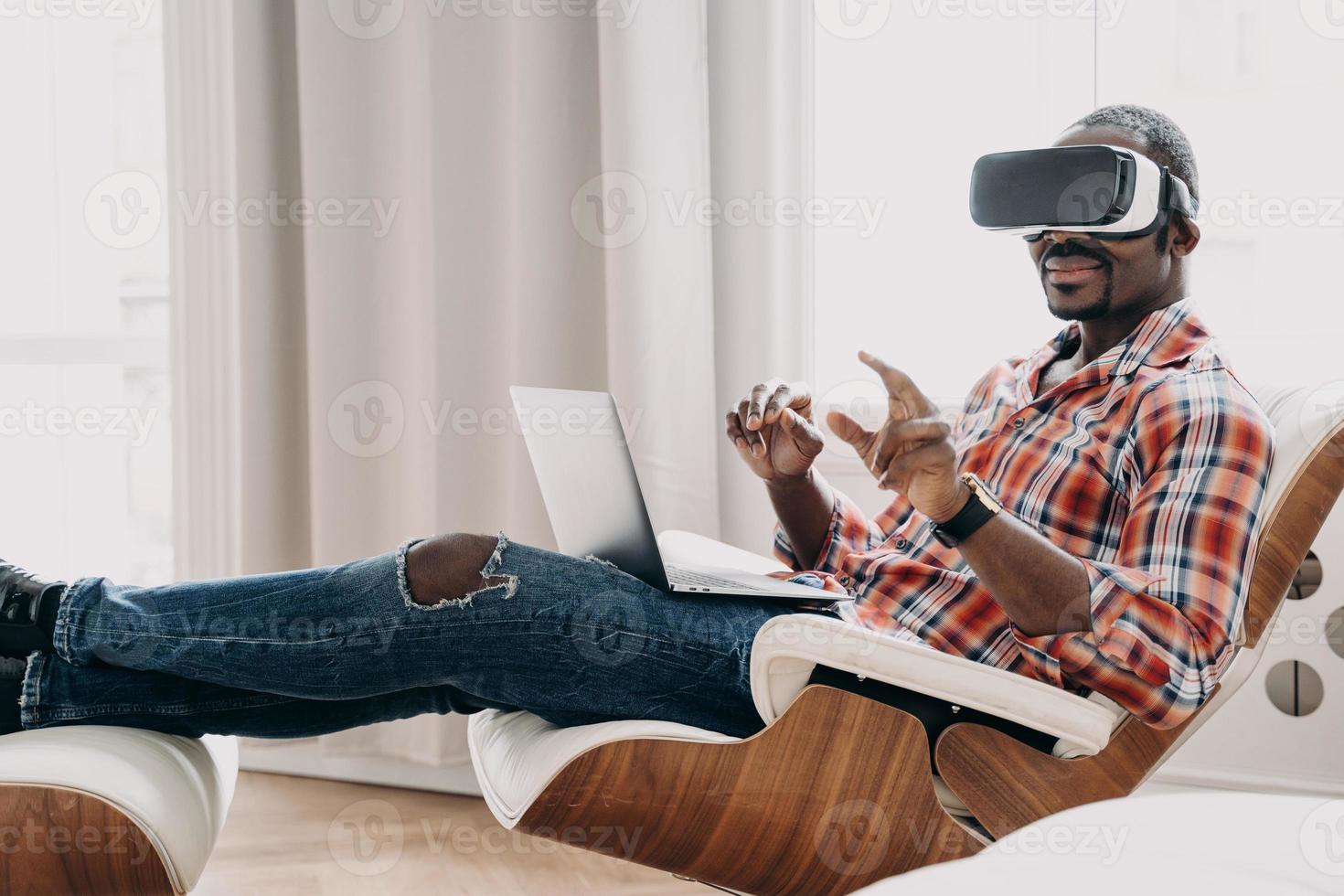 African american man in VR glasses making purchases in augmented reality store at laptop. E commerce photo