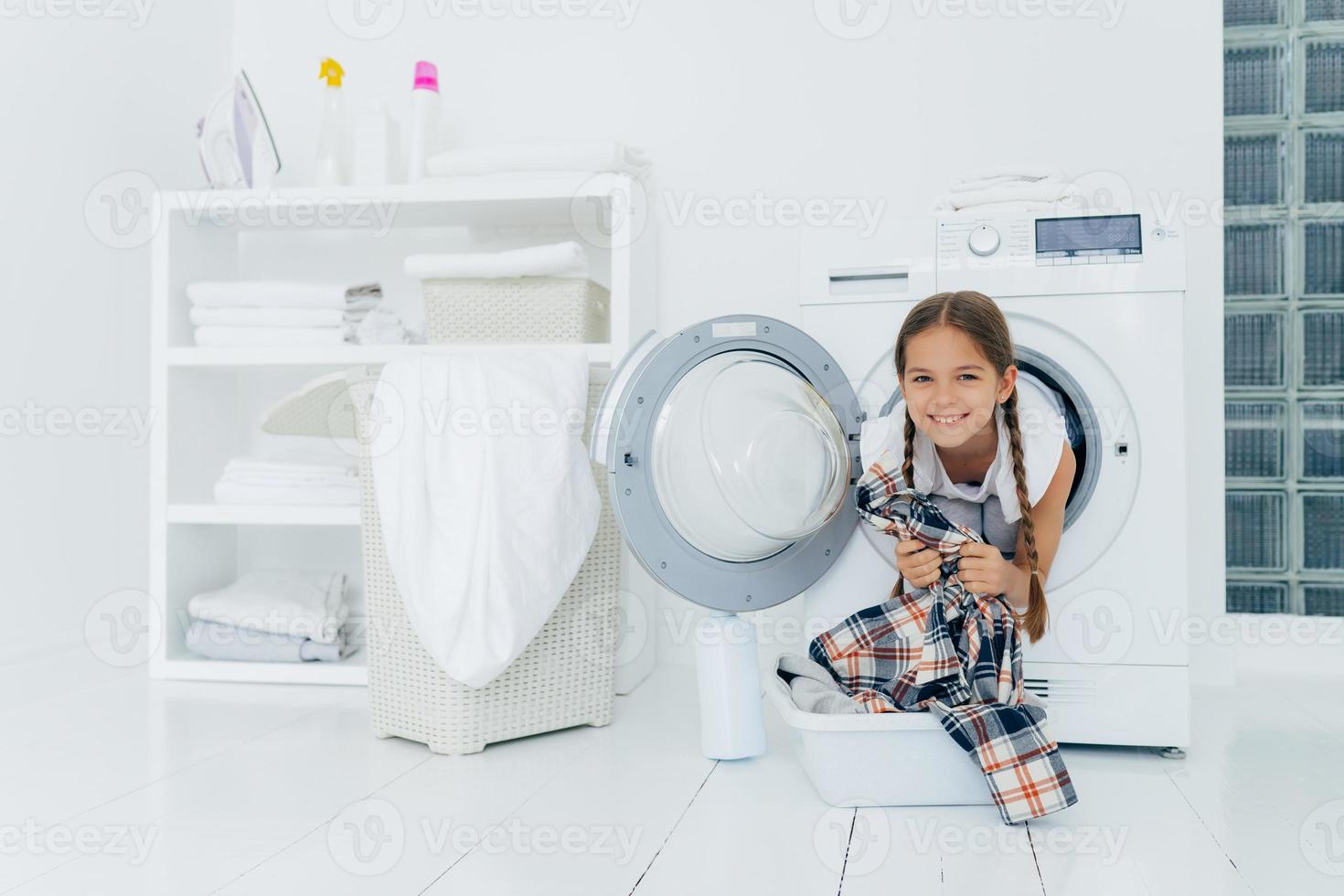 Positive kid with pigtails sticks out head from washing machine, has fun and holds shirt, prepares for washing, smiles gently, spends free time in laundry room. Children, cleanliness, hygiene concept photo