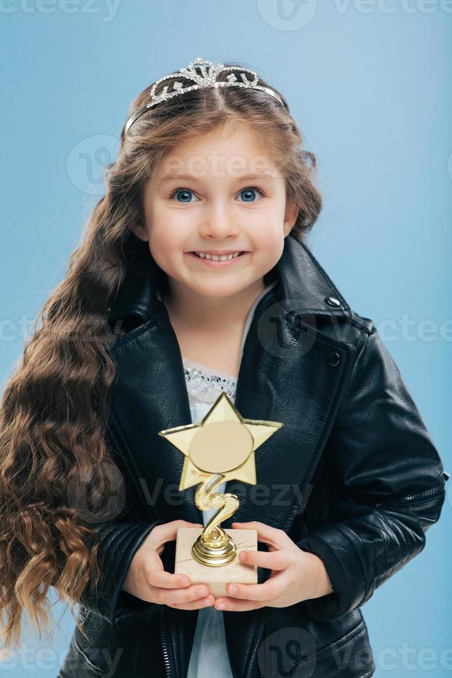 niño europeo sonriente positivo con ojos azules, cabello largo y crujiente, tiene recompensa en forma de estrella, usa chaqueta de cuero negro, aislado sobre fondo azul. la imagen de una chica feliz posa en el interior tiene buen humor foto