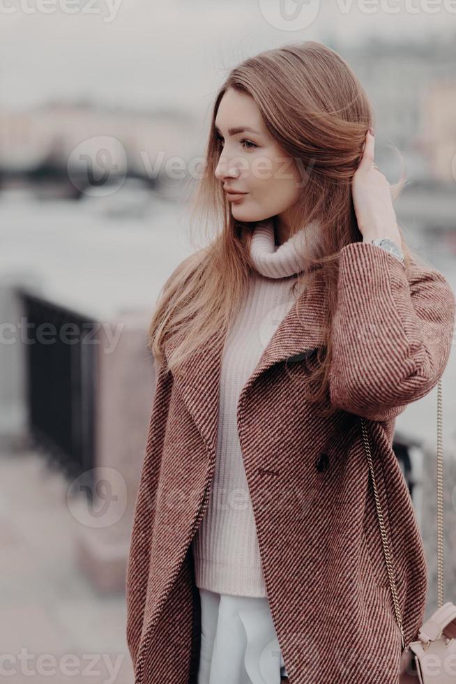 Vertical shot of fashionable Caucasain woman looks aside, wears fashionable warm coat, poses outdoor, enjoys day off, has date. Beautifu young lady goes to work, carries womans bag. Urban style photo