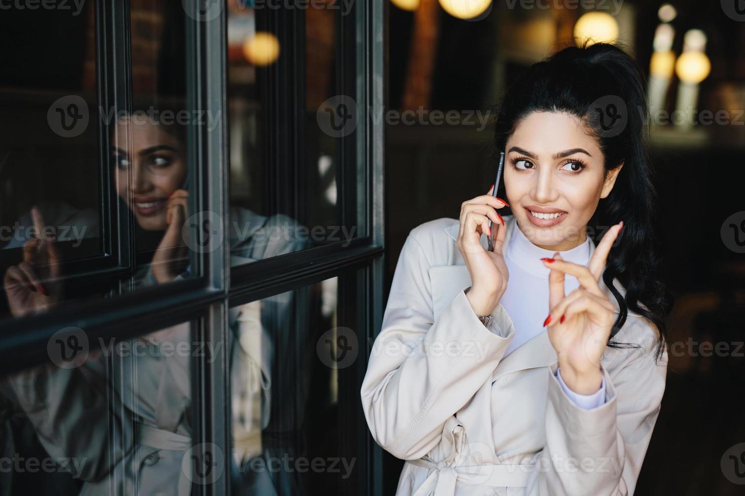 encantadora joven morena con una sonrisa agradable con manos suaves y hermosa manicura comunicándose por teléfono móvil con su amiga levantando el dedo mientras tiene una buena idea para la fiesta foto