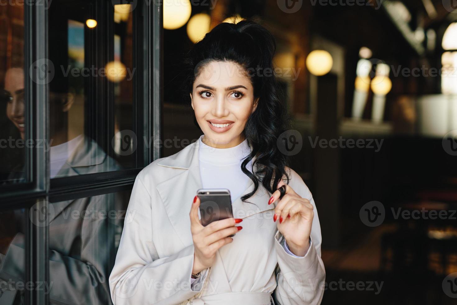 retrato horizontal de una adorable mujer morena vestida de blanco usando su teléfono celular mientras está de pie afuera con buen humor tocando su rizo negro de cola de caballo tratando de tomar una decisión. niña elegante foto