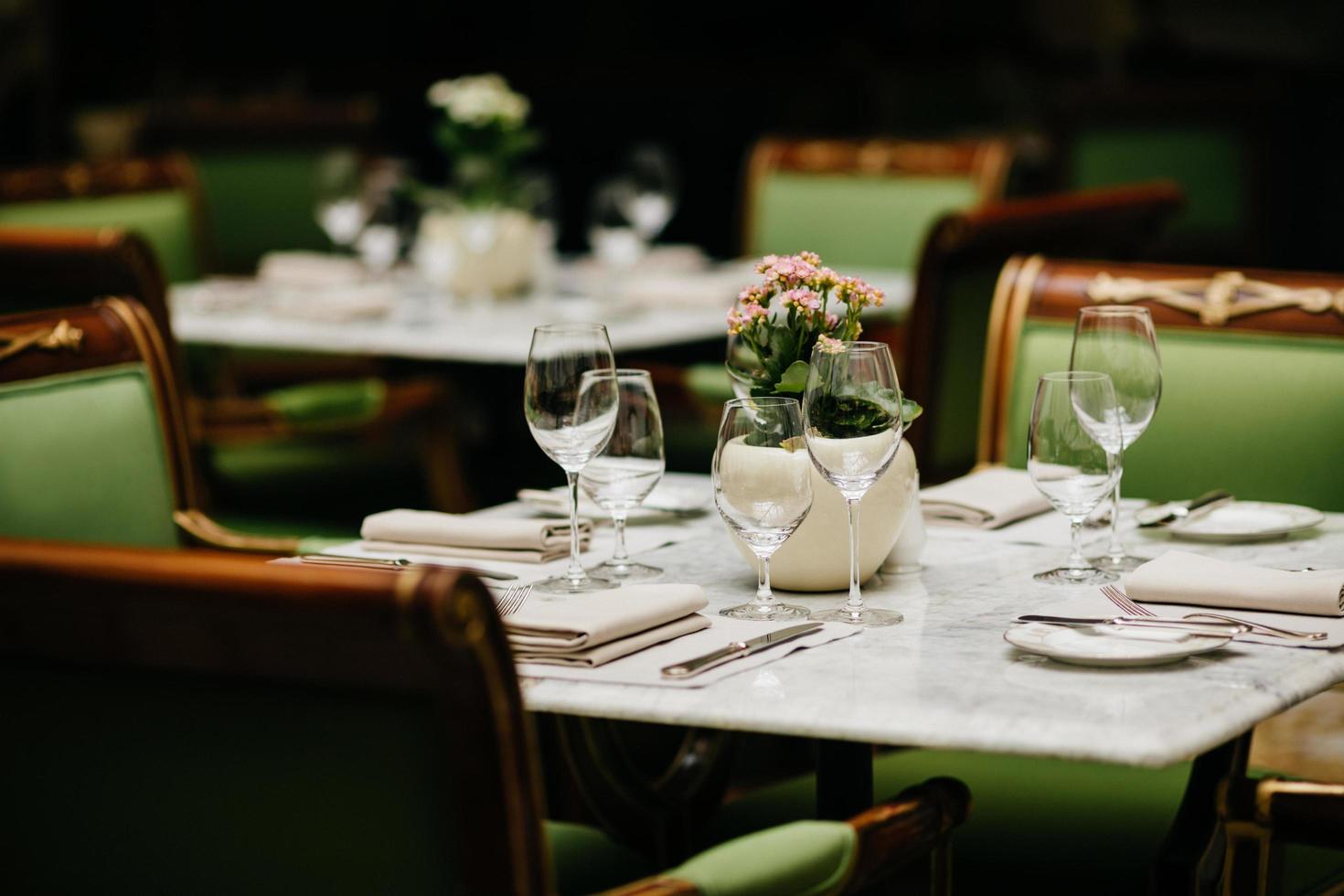 Horizontal shot of square table served with empty glasses, napkins, plates, forks and knives with no dish, green chairs around. Cozy restaurant or cafe photo