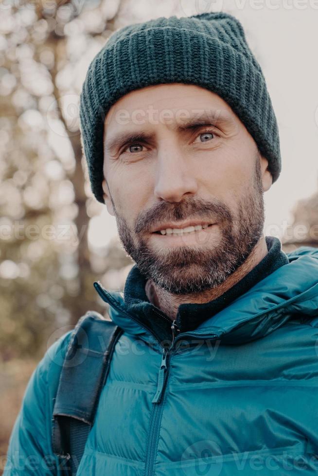 el primer plano del hombre de ojos azules tiene cerdas oscuras, mira seriamente a la cámara, usa sombrero y chaqueta, posa afuera, camina al aire libre, tiene una apariencia agradable. gente, estilo de vida, concepto de masculinidad foto