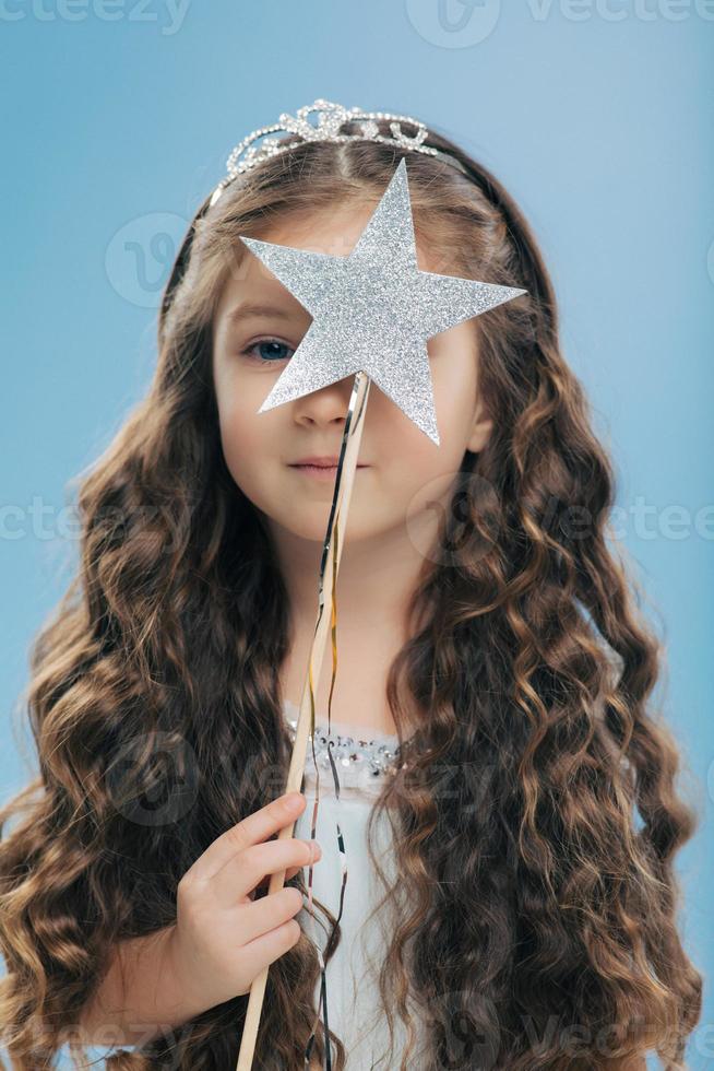 Indoor shot of good looking small female child with long curly hair, wears crown, covers face with magic wand in form of star, isolated over light blue background. Childern and magic concept photo