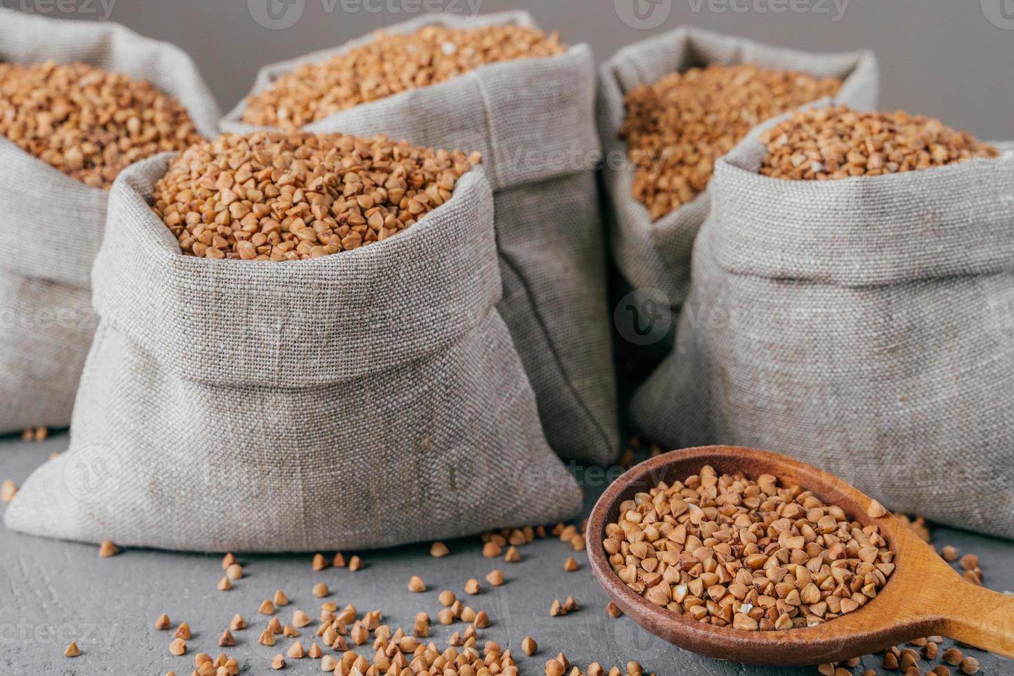 Close up shot of little sack made of linen fabric filled with dry brown buckwheat, wooden spoon with raw cereal. Selective focus photo