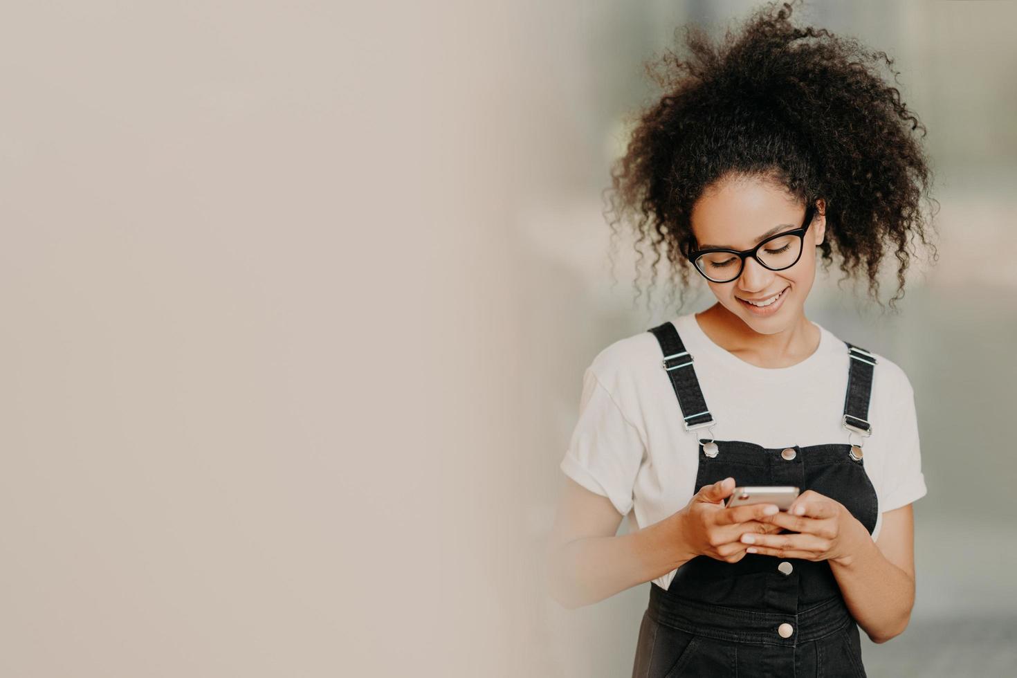 Beautiful African American girl in fashionable clothing, uses mobile phone for sending text messages, connected to wifi, checks email box, stands near white wall with empty space. Online communication photo