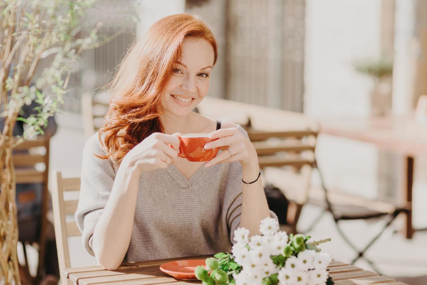Horizontal shot of happy red haired woman holds cup of drink, with pleased expression, has flowers on table, enjoys date with boyfriend, sunny day, has coffee break in cosy outdoor cafeteria. photo