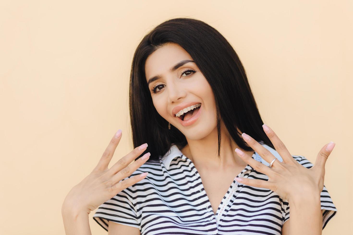 Hey look at me Beautiful brunette young female with healthy skin, make up, smiles joyfully at camera, wears braces on teeth, keeps hands in front of herself, being in good mood. Beauty concept photo