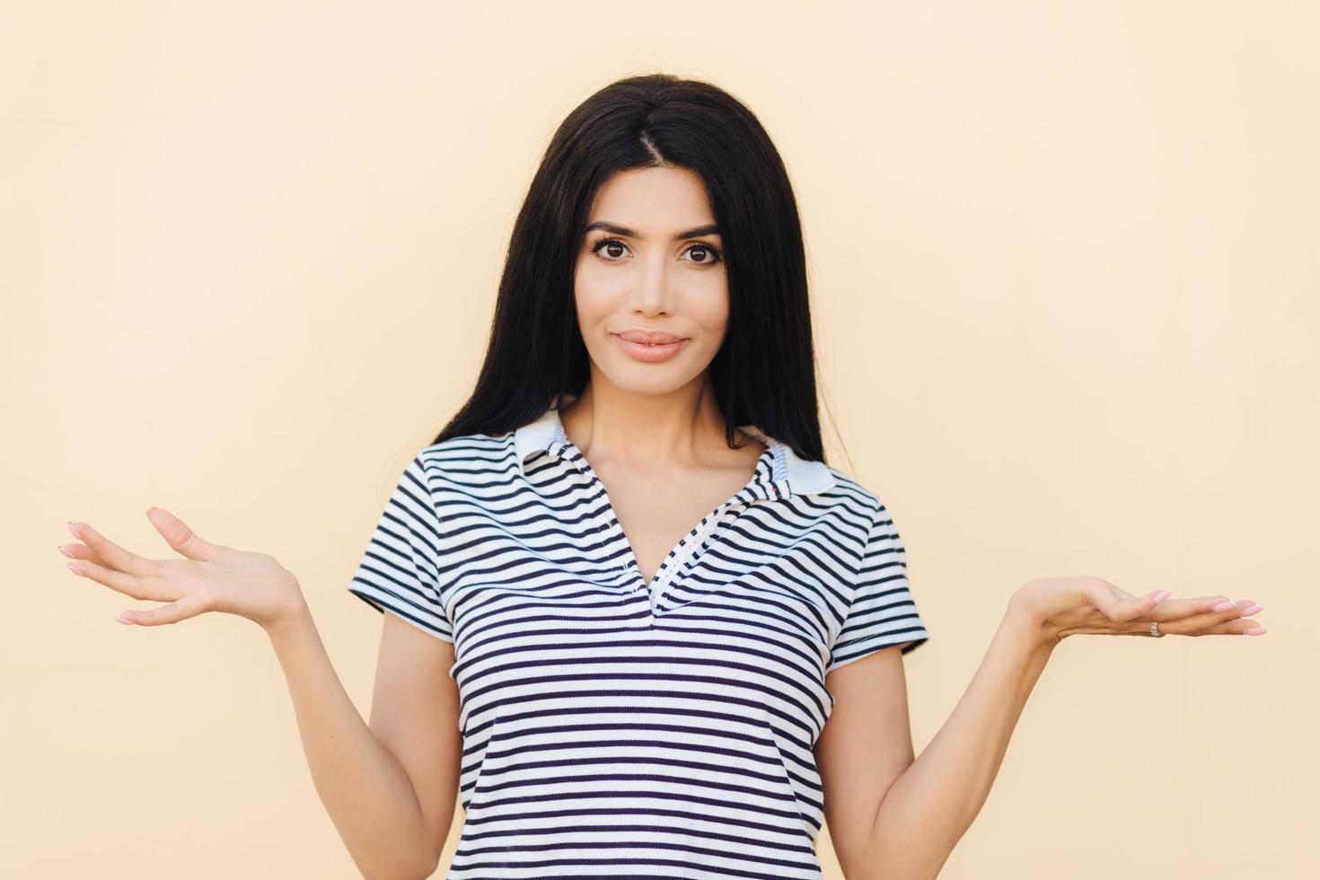 Who cares Adorable brunette young female with hesitant expression, shrugs shoulders in bewilderment, tries to find solution or makes decision, poses indoor alone. People and hesitation concept photo