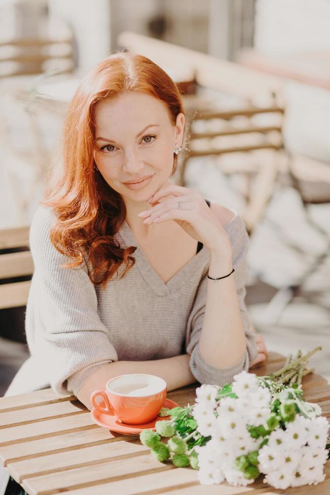 concepto de personas y estilo de vida. la mujer pelirroja tiene una expresión complacida, bebe café, posa en la cafetería con flores, tiene una cita con su novio, mira directamente a la cámara, disfruta del tiempo libre en la terraza foto