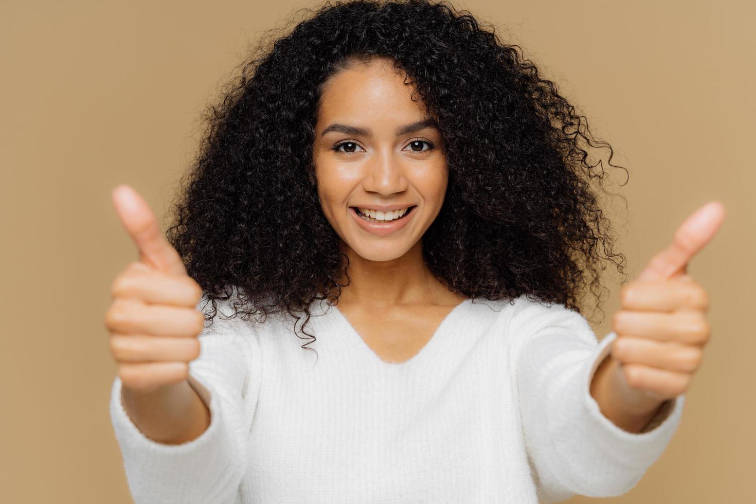 Portrait of good looking female raises thumbs, shows like gesture in camera, enjoys nice presentation, dressed in casual clothes, poses against brown background. Approval and recommendation concept photo